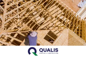 A person in a blue shirt and hat works on the wooden frame structure of a building's roof under construction, meticulously handling the roofing materials. The Qualis Roofing & Construction logo is visible at the bottom.